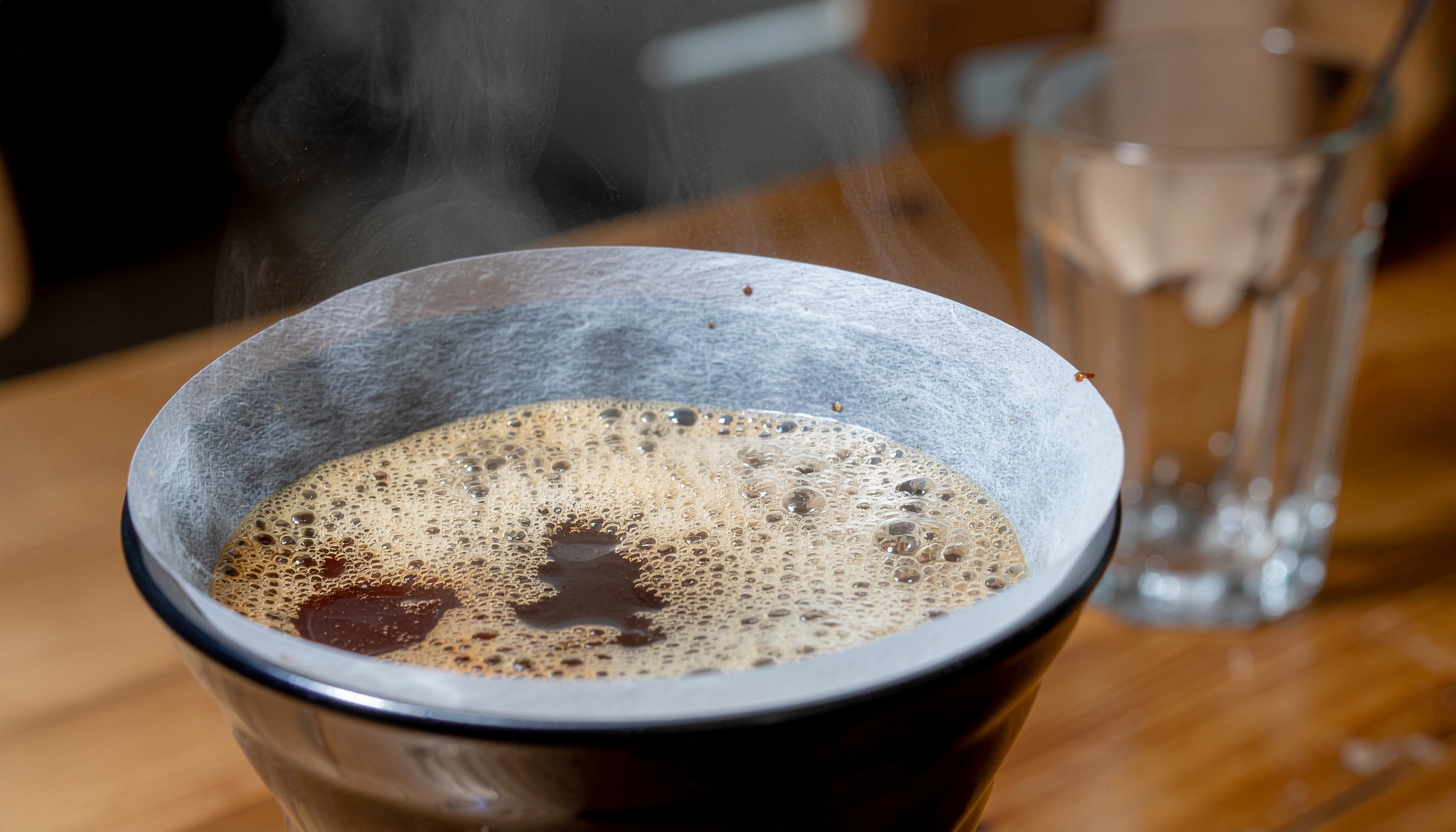 Close-up de um coador de café com borra efervescente e vapor visível, sobre uma mesa de madeira. A luz suave destaca texturas e cria um foco nítido na bebida quente. Ao fundo, um copo de vidro desfocado adiciona profundidade à composição minimalista da cena.